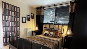 A bedroom with a metal-framed bed, a bookshelf, a radiator, and a window with blinds.