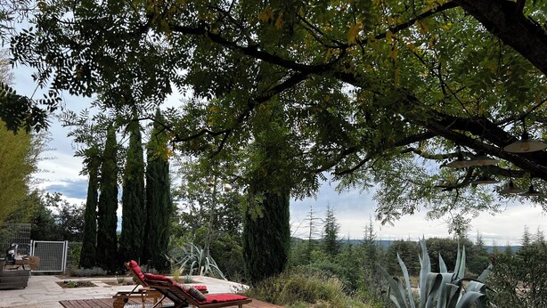 A wooden deck with a lounge chair and a table, surrounded by tall trees and a view of the landscape.