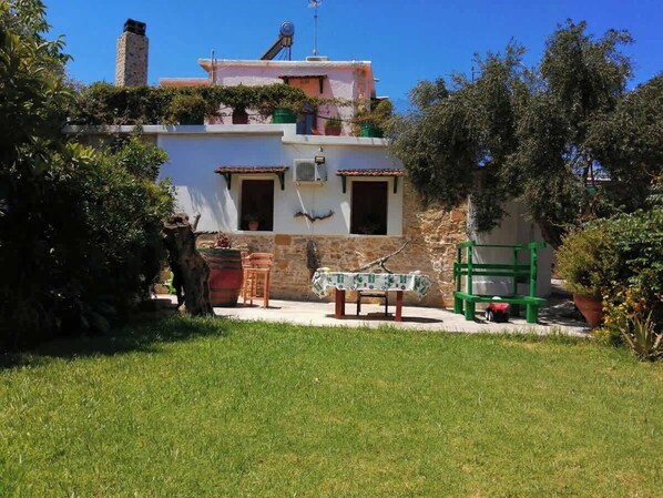 Una casa de dos pisos con un balcón, una pequeña mesa con sillas y un banco verde.