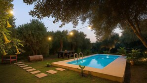 Una piscina rectangular con interior azul claro, rodeada por un césped con piedras que forman un sendero hacia un banco y una mesa de madera.