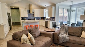 A modern kitchen with white cabinets, a central island, and orange stools. There is a brown sofa with a throw blanket and a colorful rug.