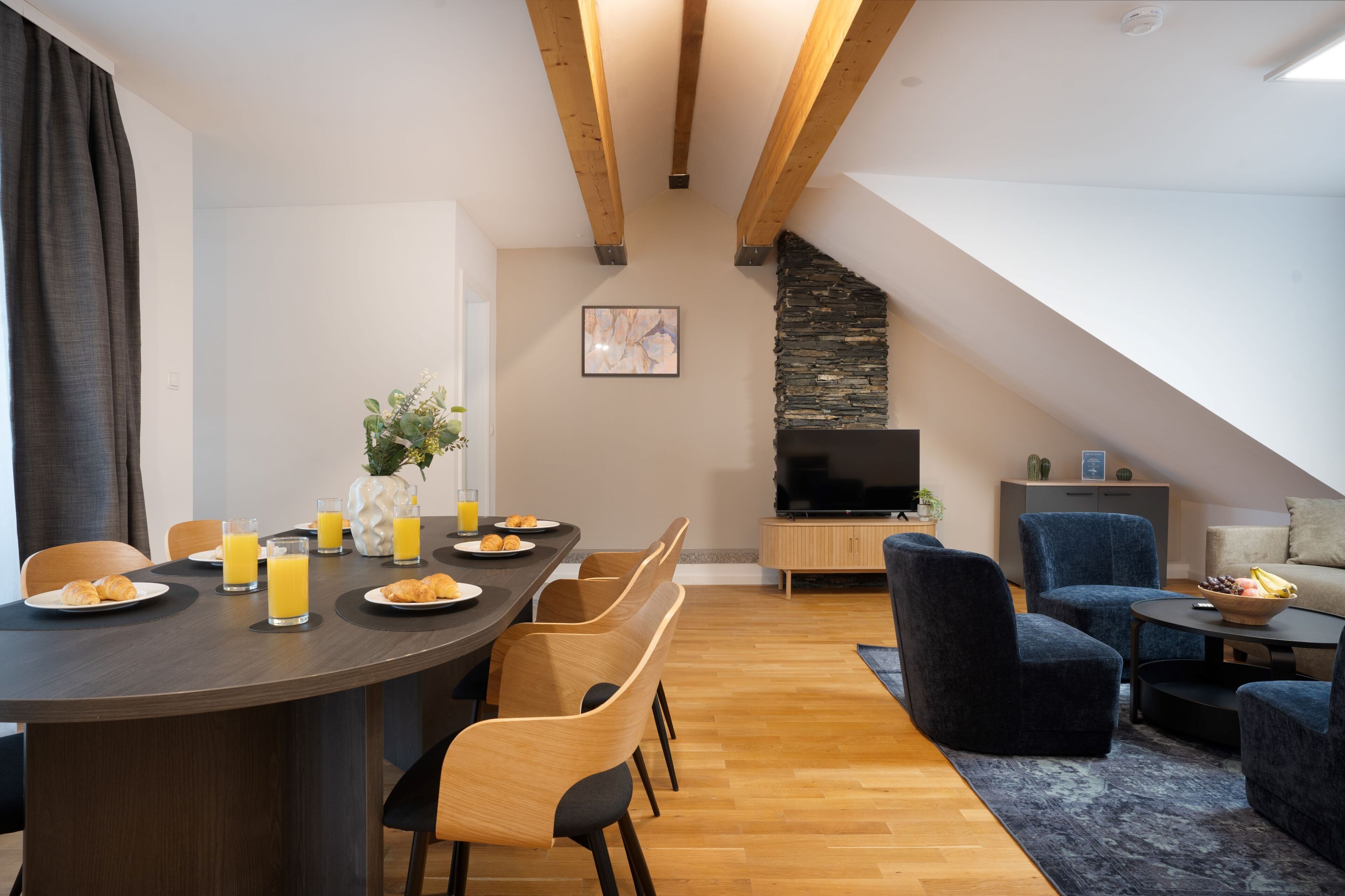 Un coin repas moderne avec une table en bois, des chaises et des éléments pour le petit-déjeuner. On aperçoit une cheminée en pierre et un téléviseur au fond.