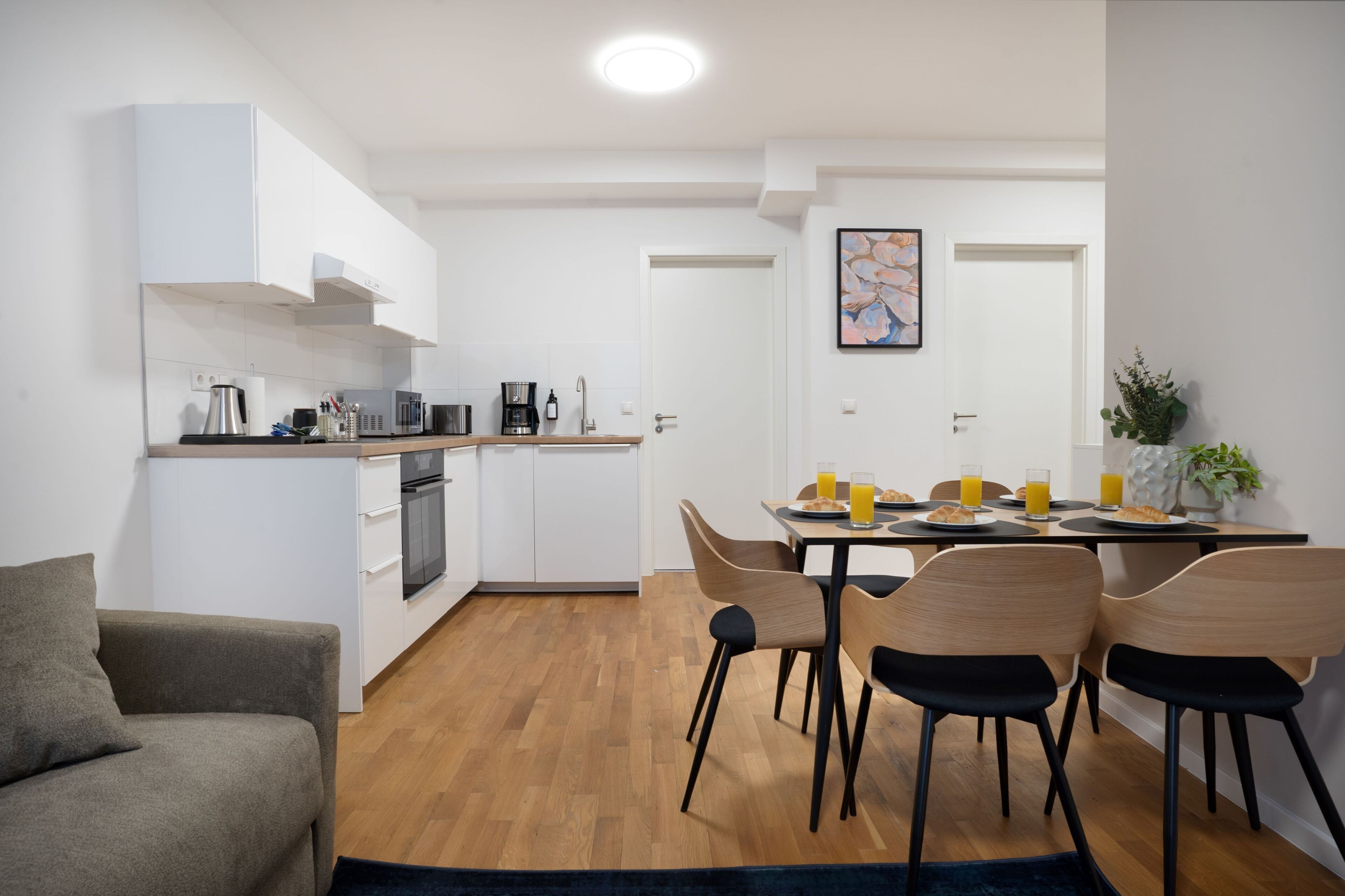 Una cocina moderna con armarios blancos, un comedor con mesa dispuesta para cuatro personas y una sala de estar con sofá.