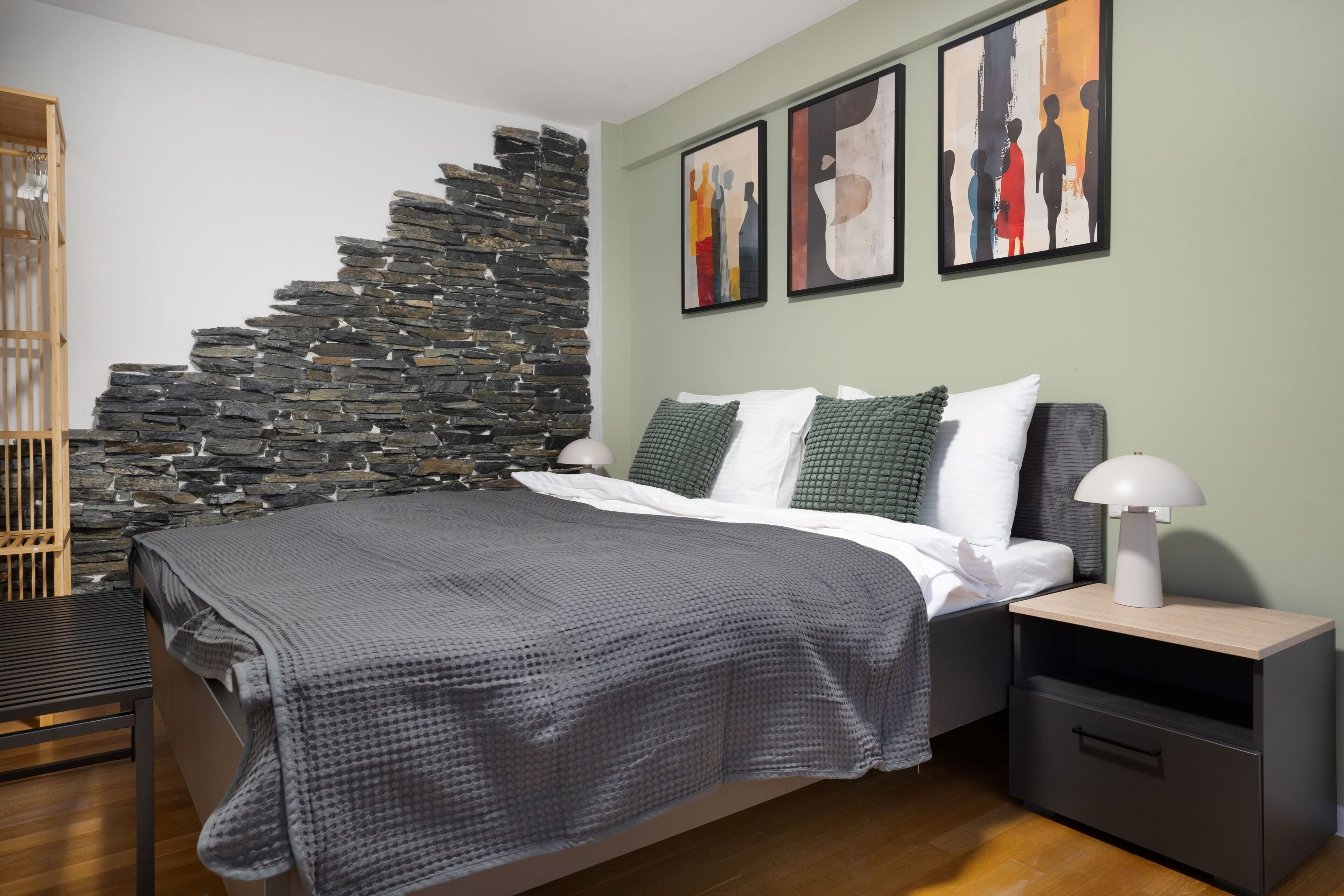 Ein Schlafzimmer mit Steinwand, einem Bett mit grauer Decke, grünen Kissen und zwei abstrakten Gemälden an der Wand.