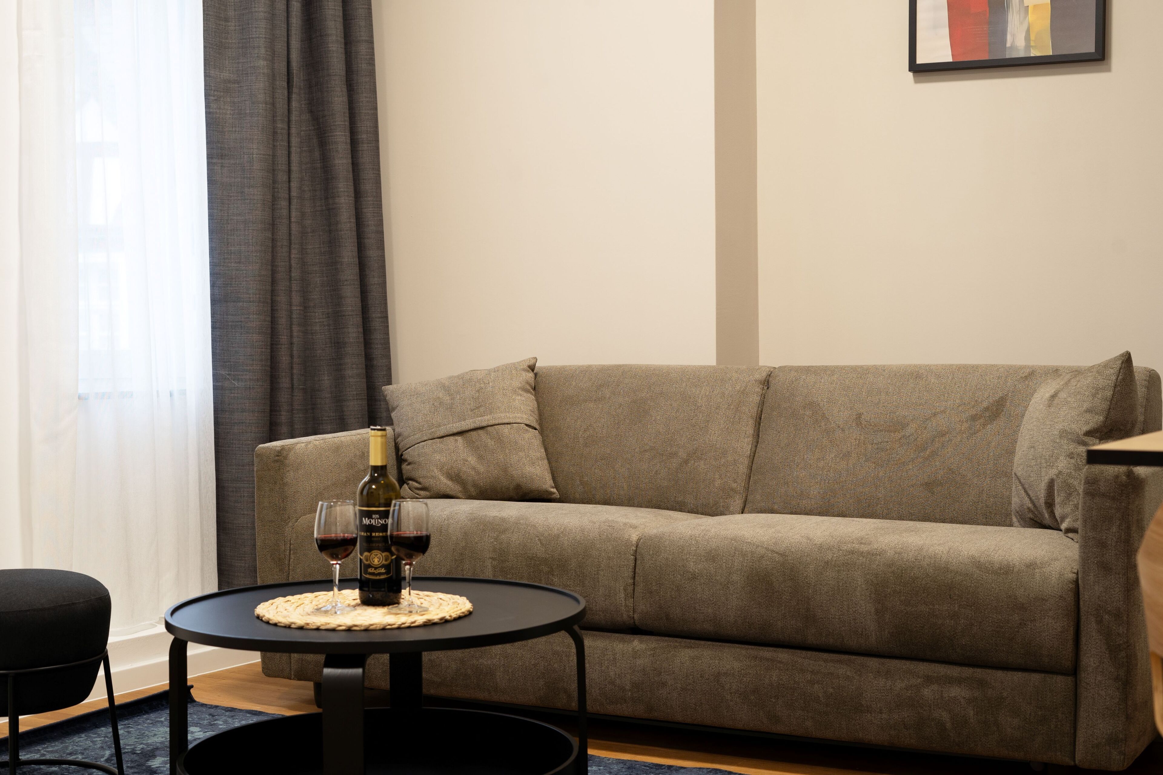 Ein Wohnzimmer mit einem beigefarbenen Sofa, einem kleinen runden Tisch mit einer Weinflasche und einem Glas sowie einem schwarzen Sessel.