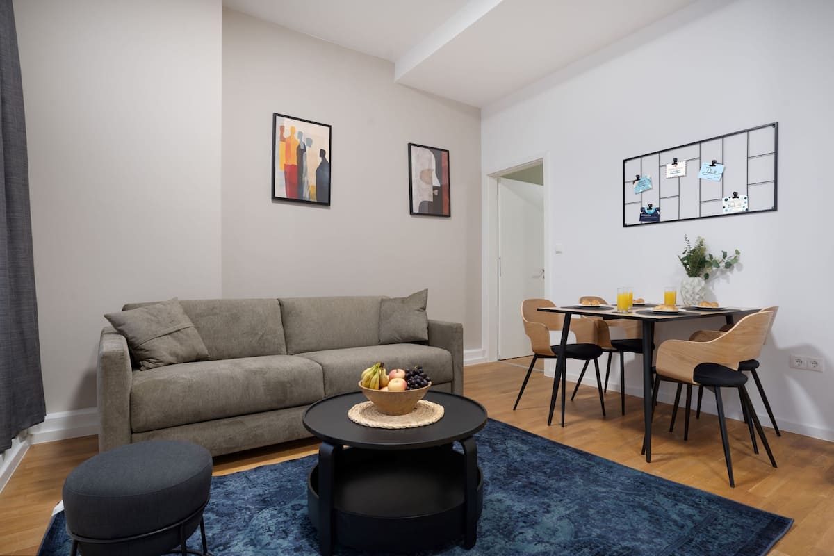 A modern living room with a grey sofa, a round coffee table with a fruit bowl, a dining table with chairs, and artwork on the walls.