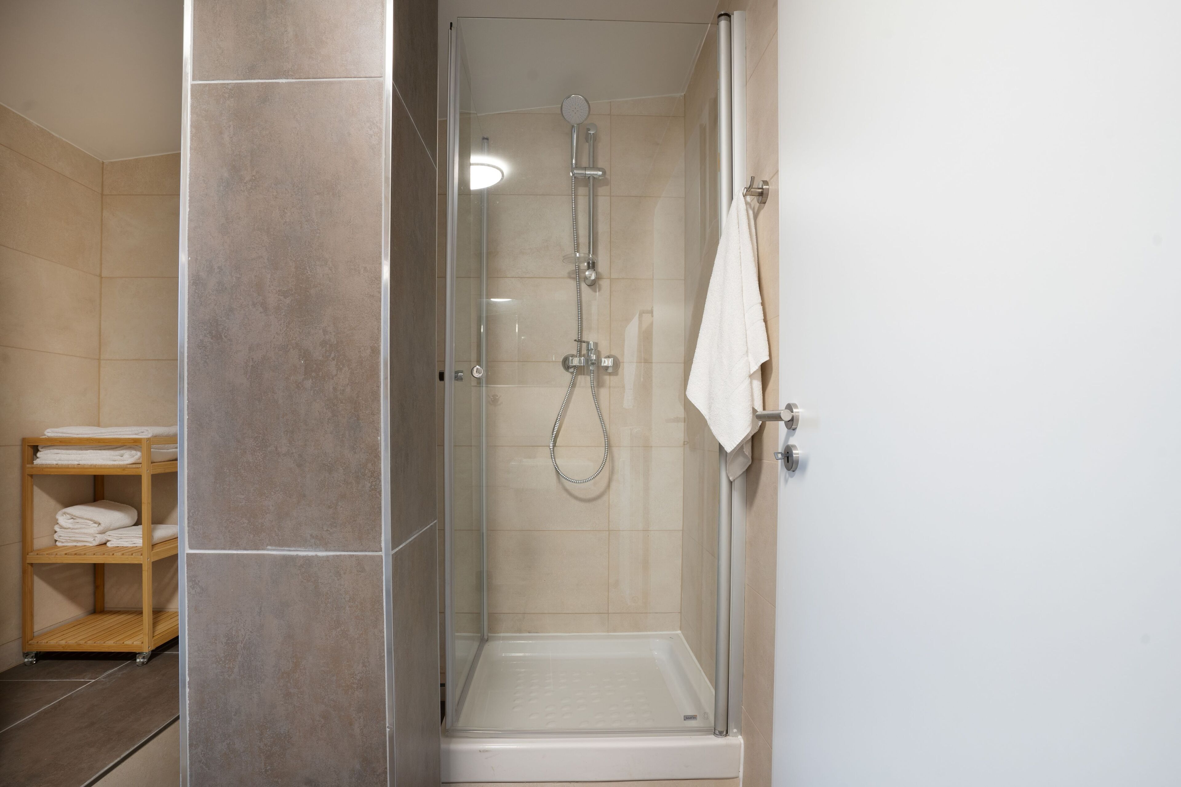 Ducha envuelta en vidrio con cabezal de ducha manual y llave de pared. Hay una toalla blanca colgada en la pared y un estante de madera con toallas dobladas.