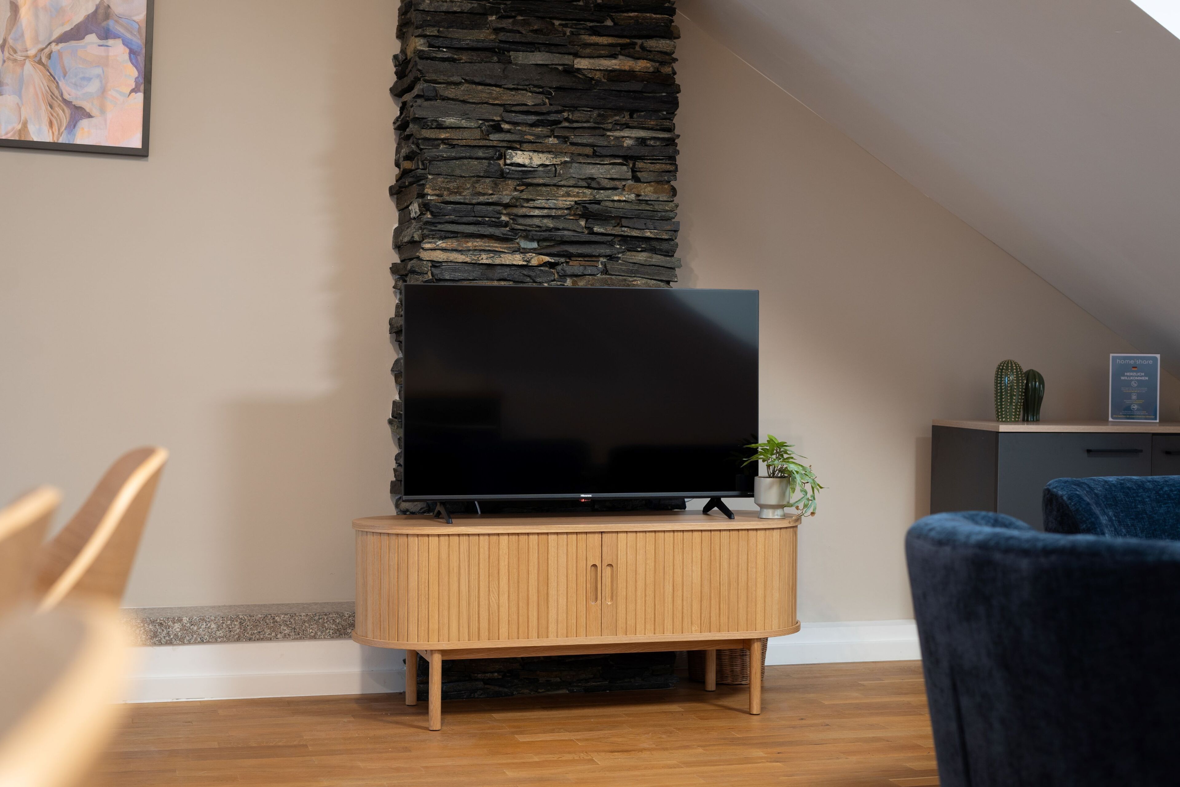 A living room with a stone wall, a flat-screen TV on a wooden cabinet, a cactus, and a blue chair.