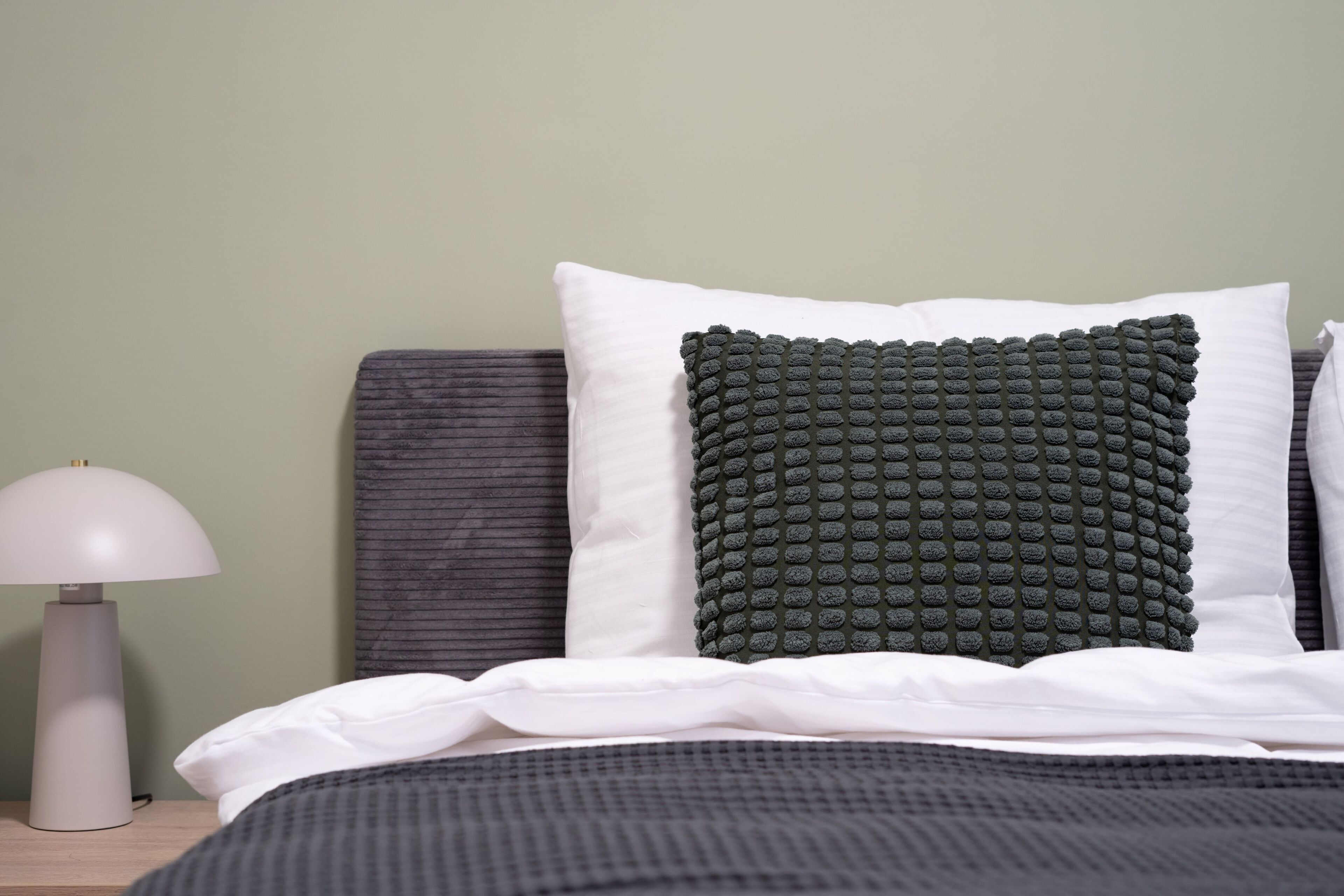 A neatly made bed with a dark headboard, white pillows, and a textured green pillow. A white lamp is on a bedside table to the left.