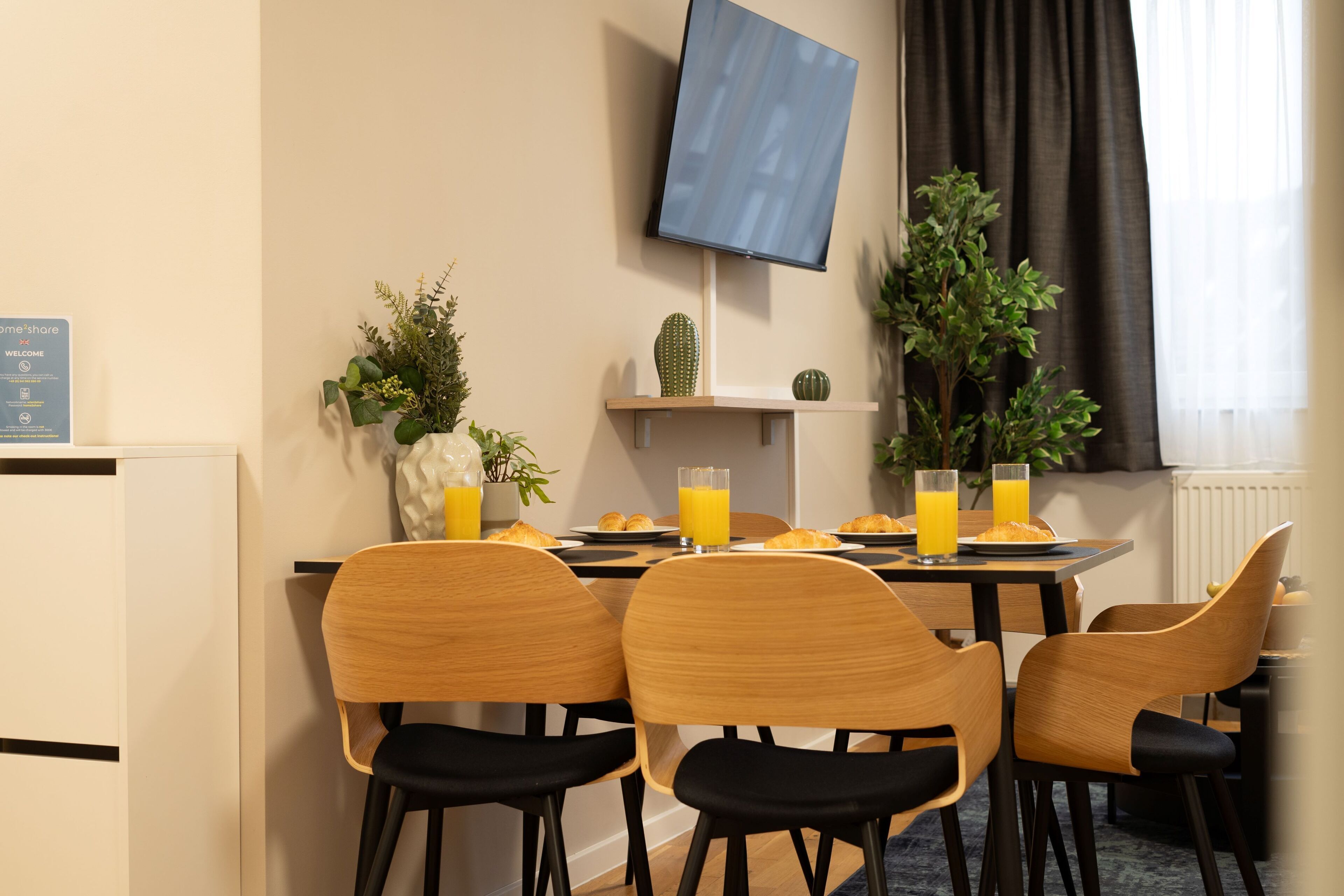 Un comedor con una mesa dispuesta para el desayuno, con croissants y zumo de naranja. Hay cuatro sillas, un televisor de pantalla plana montado en la pared y una planta en maceta sobre una estantería.