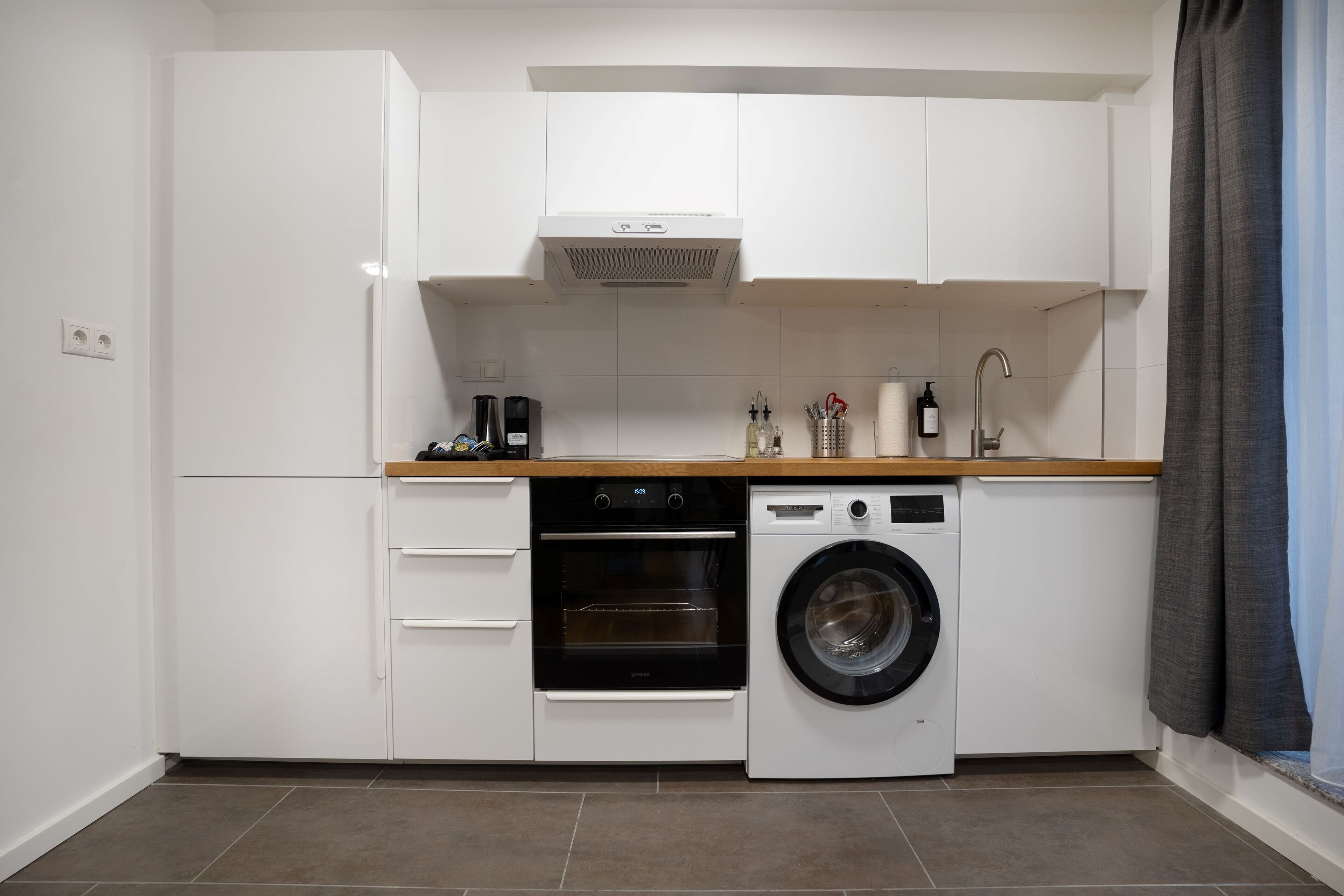 A compact kitchen with white cabinets, a built-in oven, a washing machine, and a coffee maker.