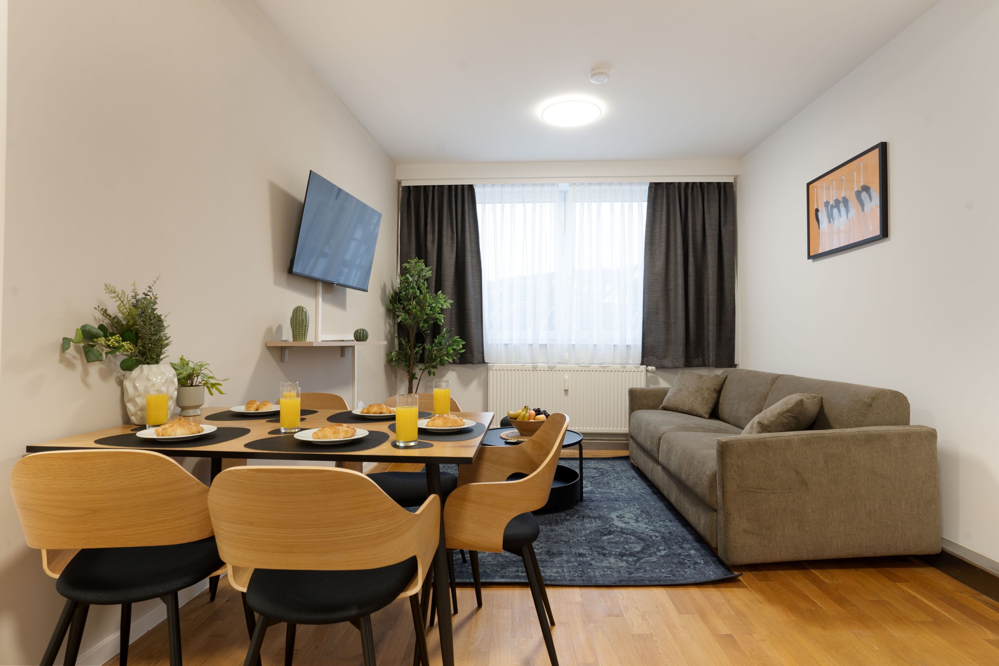 A modern living room with a sofa, a dining table with chairs, a TV mounted on the wall, and a window with curtains.