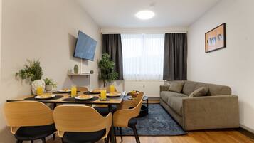 A modern living room with a sofa, a dining table with chairs, a TV mounted on the wall, and a window with curtains.