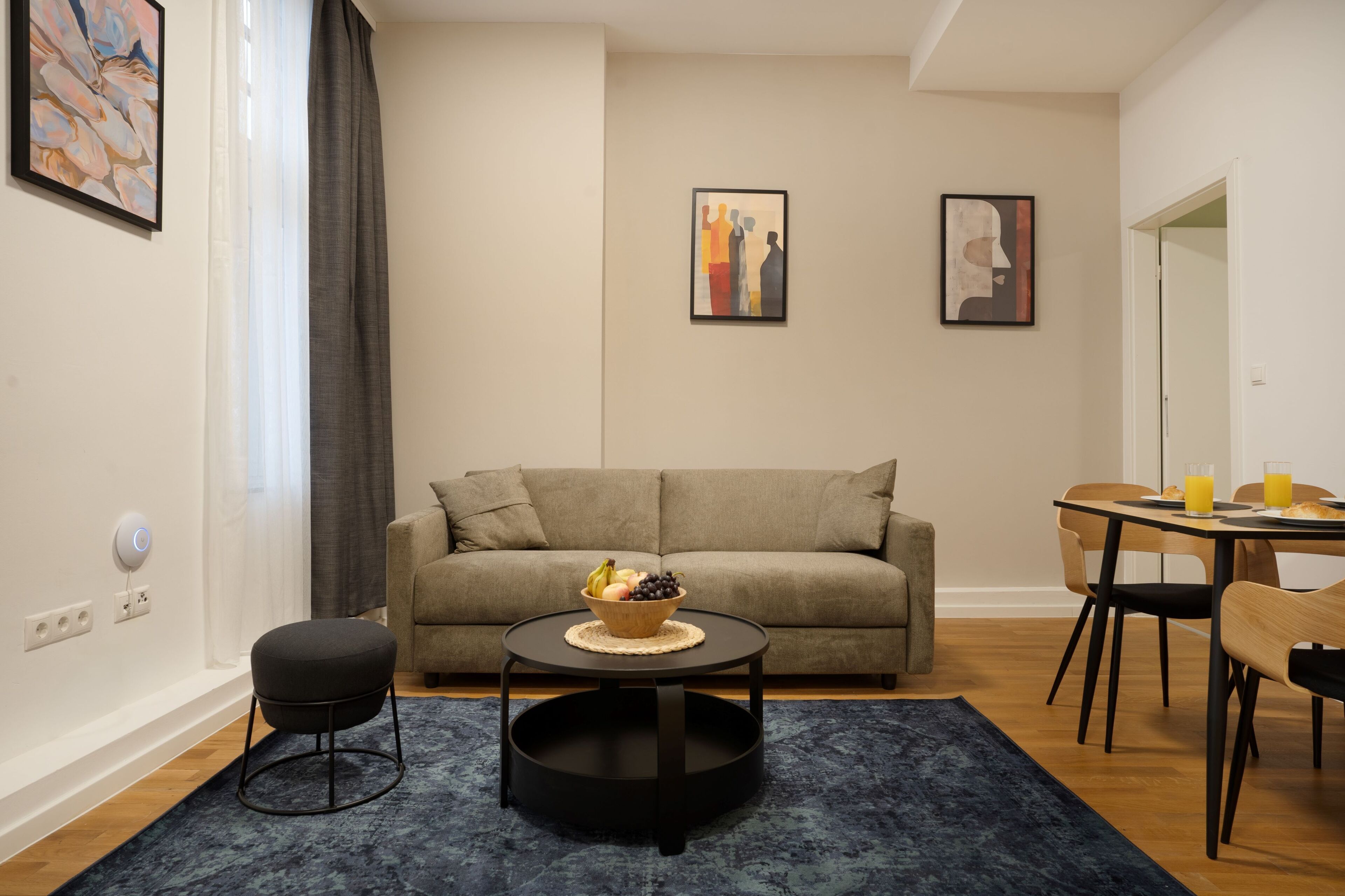 Un salon moderne avec un canapé, une table basse ronde sur laquelle sont disposés des fruits, une table à manger avec des chaises et des œuvres d’art accrochées aux murs.