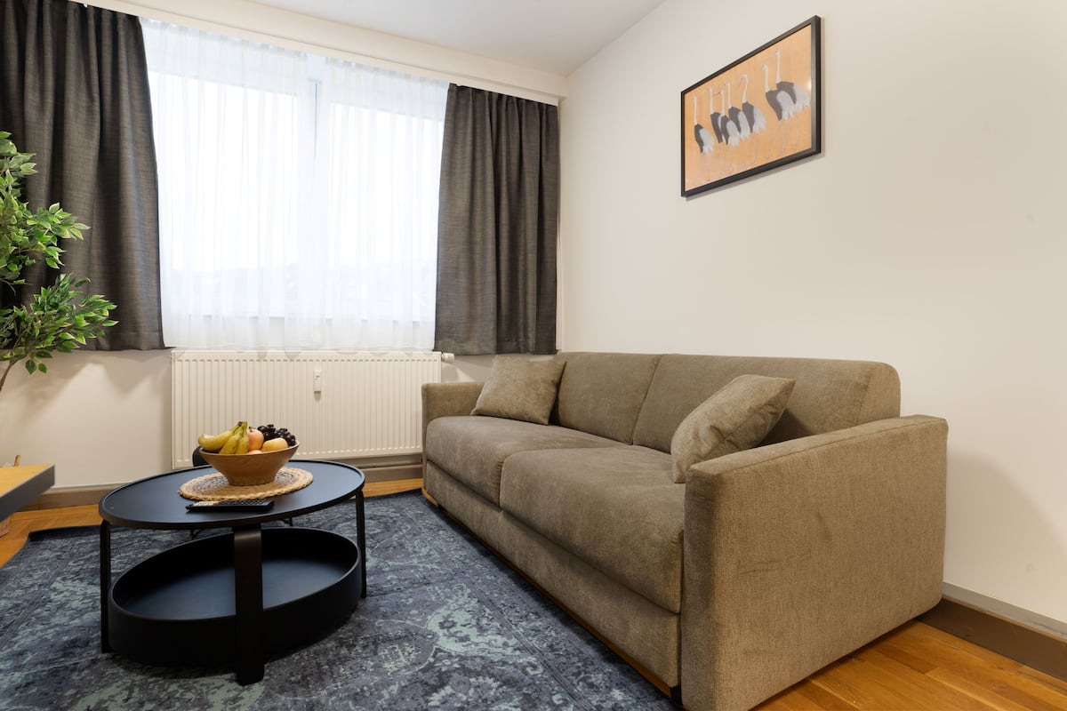 A living room with a beige sofa, a round coffee table with a bowl of fruit, a potted plant, and a framed picture on the wall.