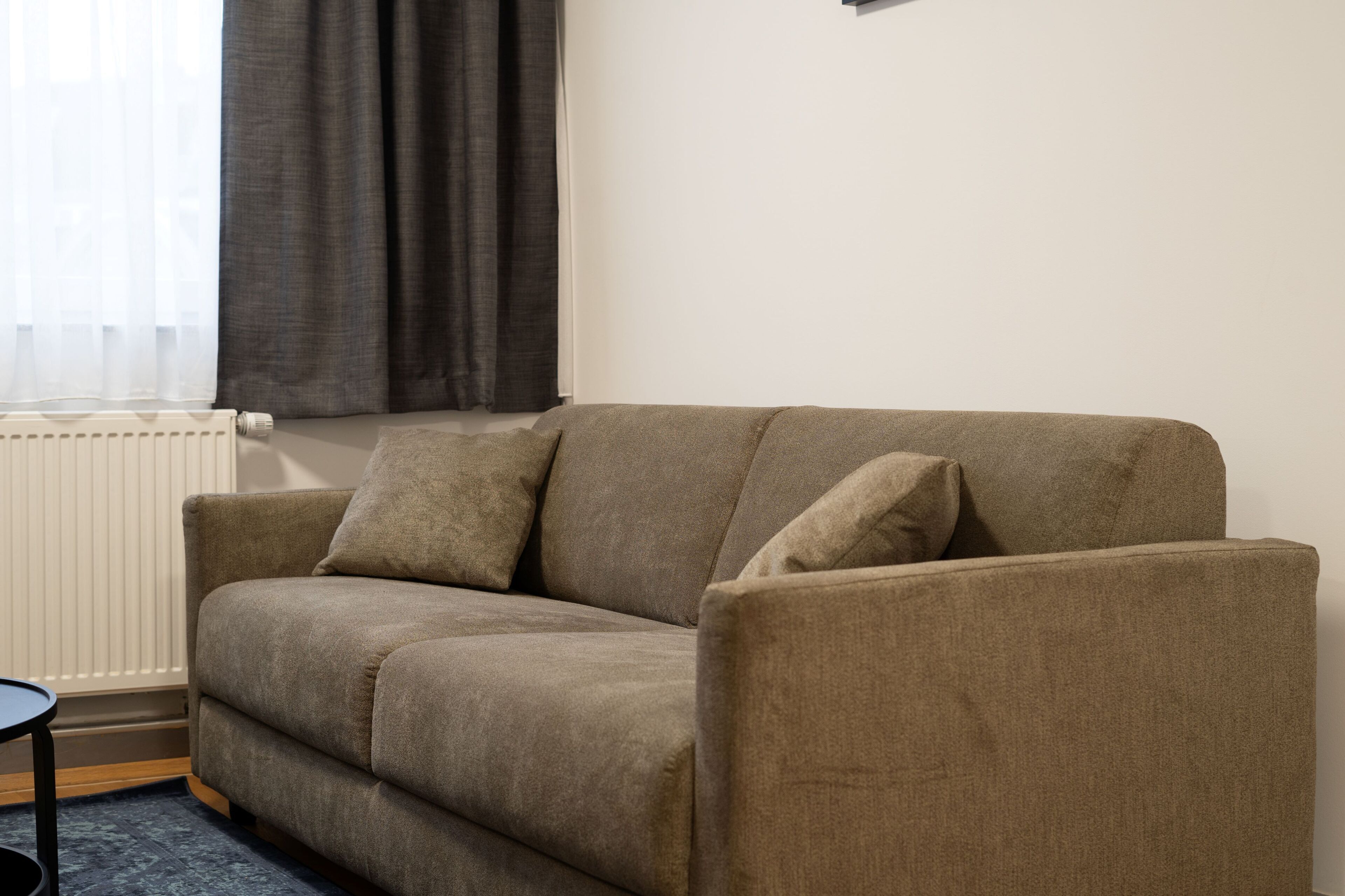 Un canapé beige avec deux coussins, dans une pièce équipée d’un radiateur et d’une fenêtre avec des rideaux.