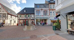 Une place pavée, bordée de bâtiments traditionnels à colombages, avec une fontaine et un magasin de vêtements.