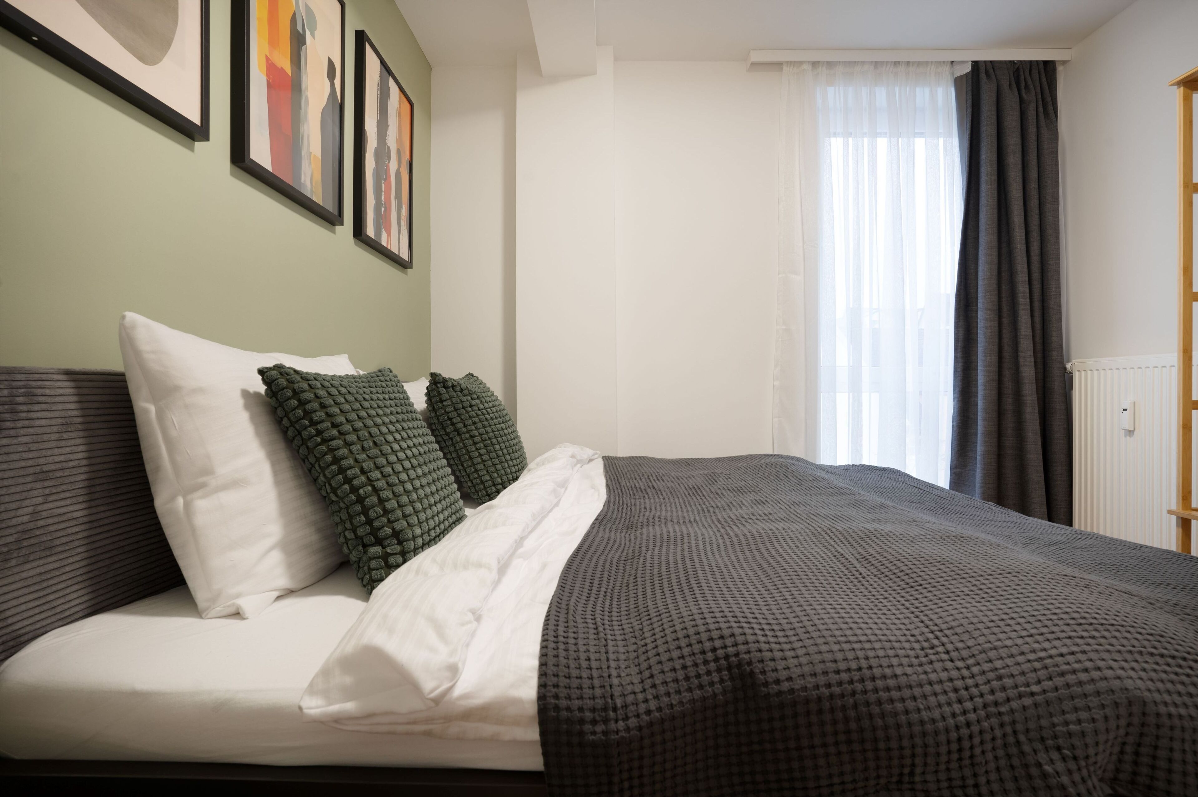 Una cama bien tendida con colcha blanca y oscura a cuadros, dos almohadas verdes y una almohada blanca. En la pared hay obras de arte enmarcadas, una ventana con cortinas traslúcidas y un radiador.