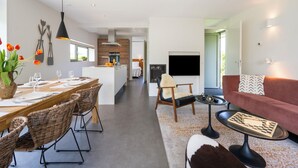 A modern living room with a sofa, a dining area with chairs, a kitchen in the background, and a TV mounted on the wall.