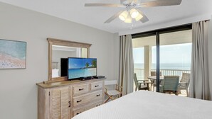 A hotel room with a bed, a television on a wooden dresser, a large window with a view of the beach, and a ceiling fan. - Summer House 901B by Liquid Life (Orange Beach)