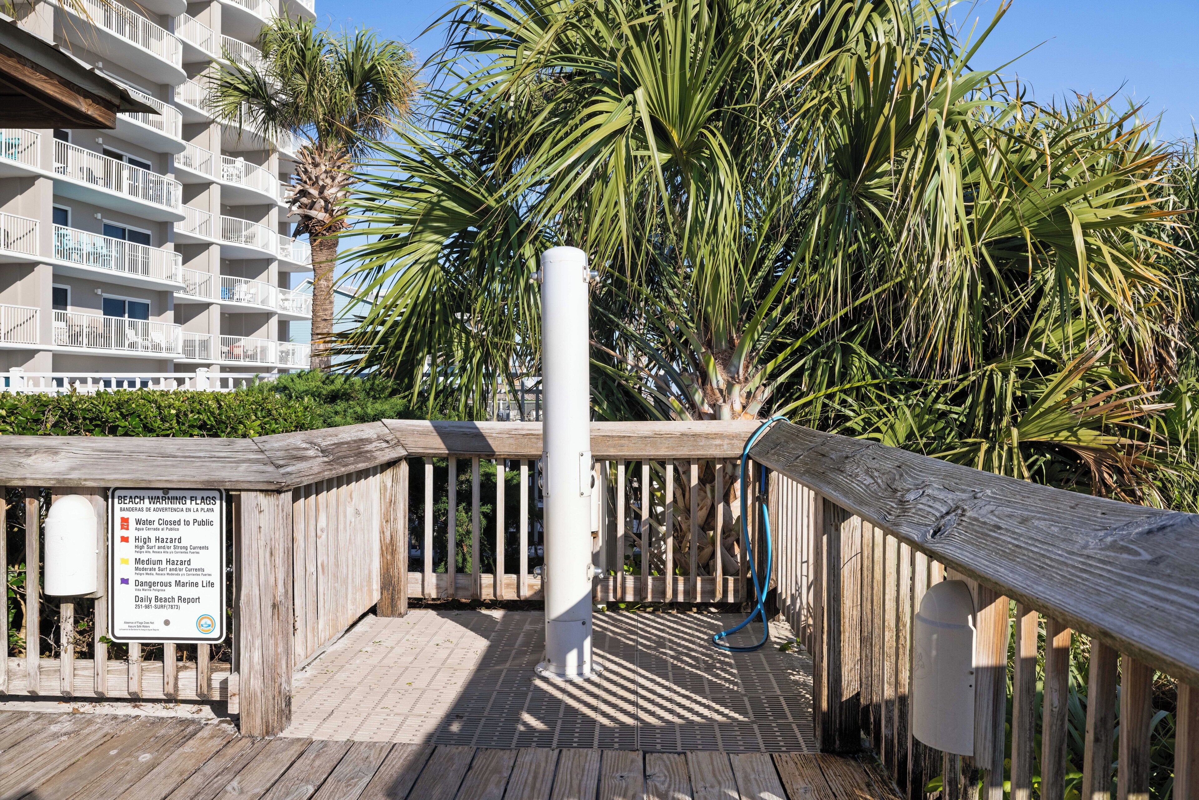 Une promenade en bois avec un panneau d’avertissement concernant la plage, des palmiers et un bâtiment en arrière-plan.