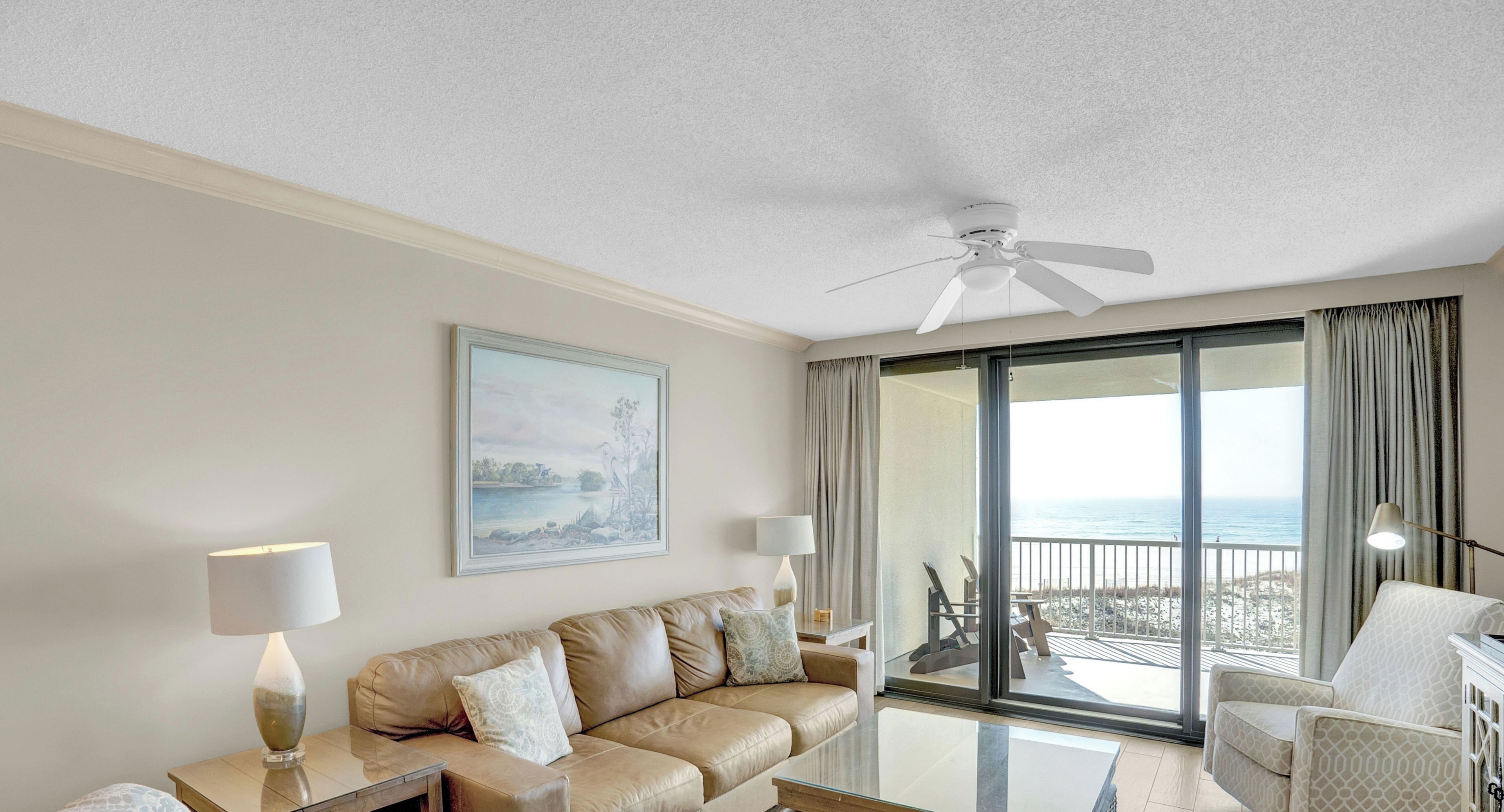 A hotel room with a sofa, a table, a lamp, a ceiling fan, and a large window with a view of the beach.