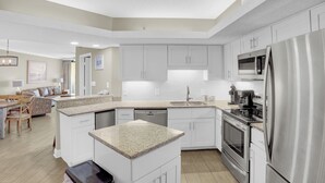 A modern kitchen with white cabinetry, a granite countertop, stainless steel appliances, and a dining area with a table and chairs. - Summer House 203B by Liquid Life (Orange Beach)