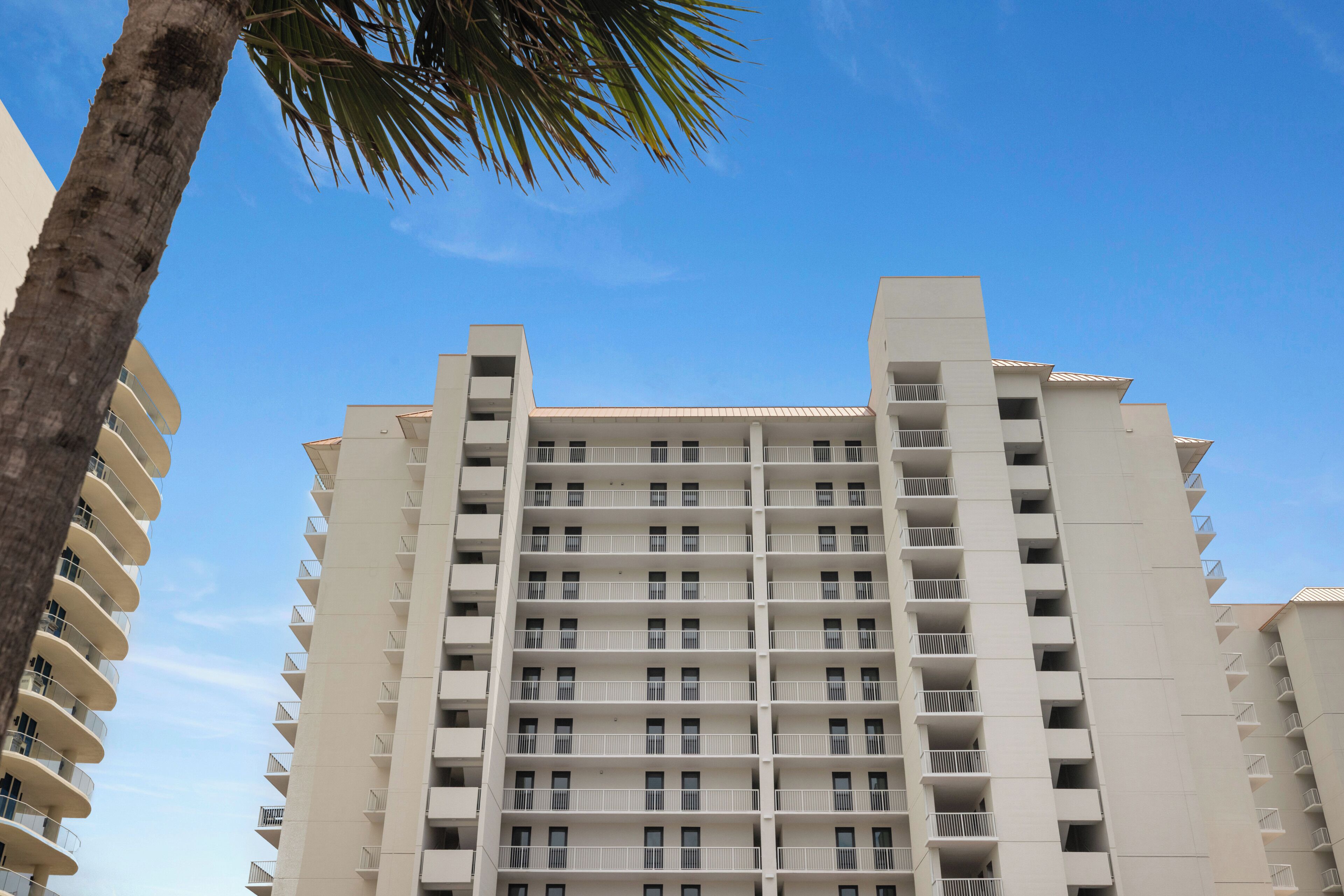 Un edificio de gran altura con varios balcones, cielo azul despejado y una palmera.