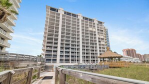 A multi-story hotel with balconies, a wooden boardwalk, and a beach area with a thatched structure. - Summer House 203B by Liquid Life (Orange Beach)