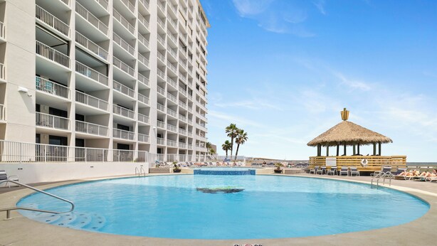 A swimming pool with a thatched-roof structure, a multi-story building with balconies, and a clear blue sky. - Summer House 203B by Liquid Life (Orange Beach)