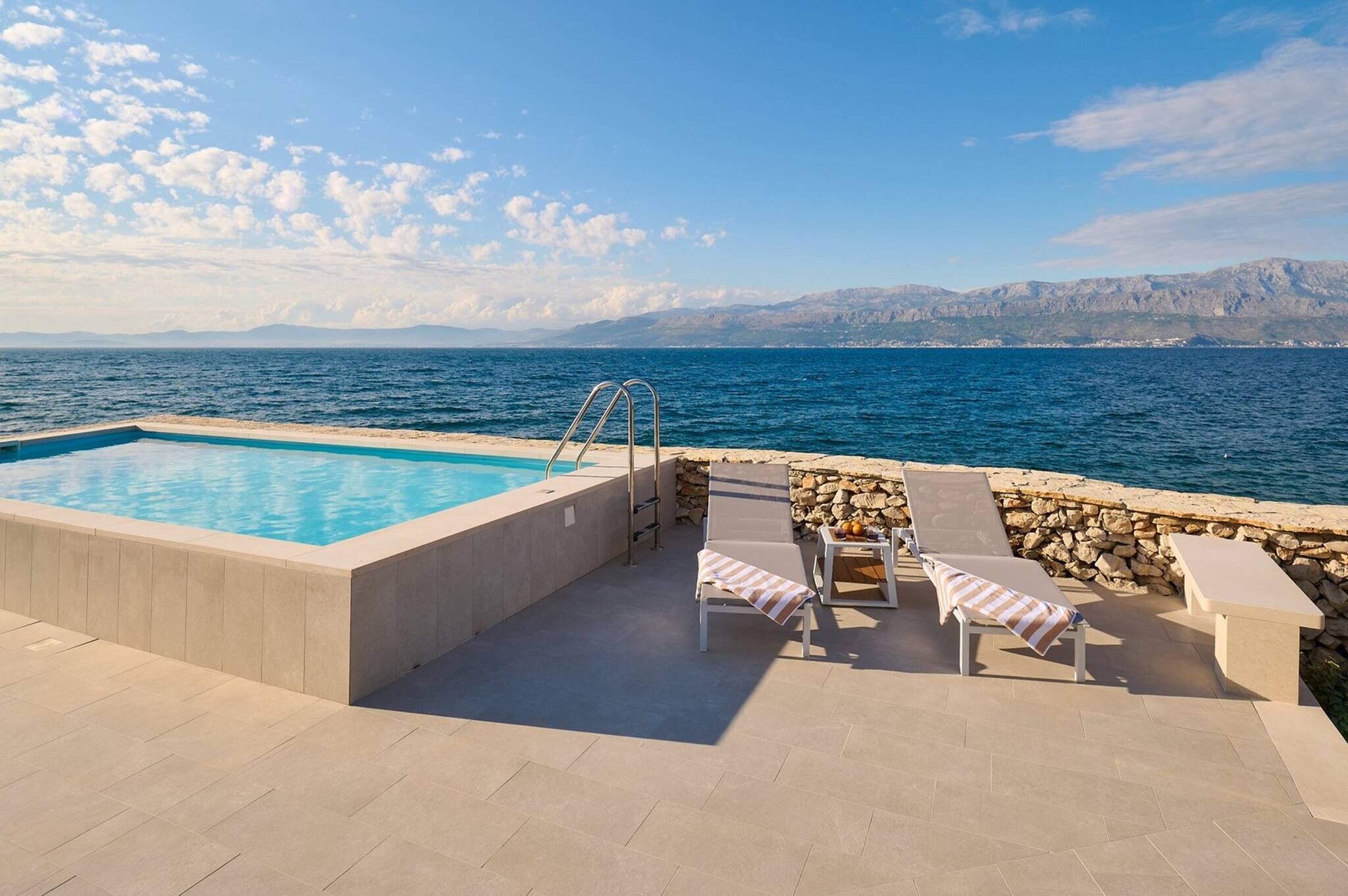 Una piscina con pared de piedra, sillas de descanso y vistas al mar y las montañas.