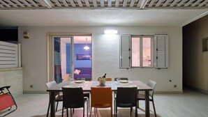 A dining area with a table set for four, a sofa in the background, and a view of a sunset through a glass door. - Ibiscus-Iblee, holiday home with garden 100 mt from the sea (Marina di Ragusa)