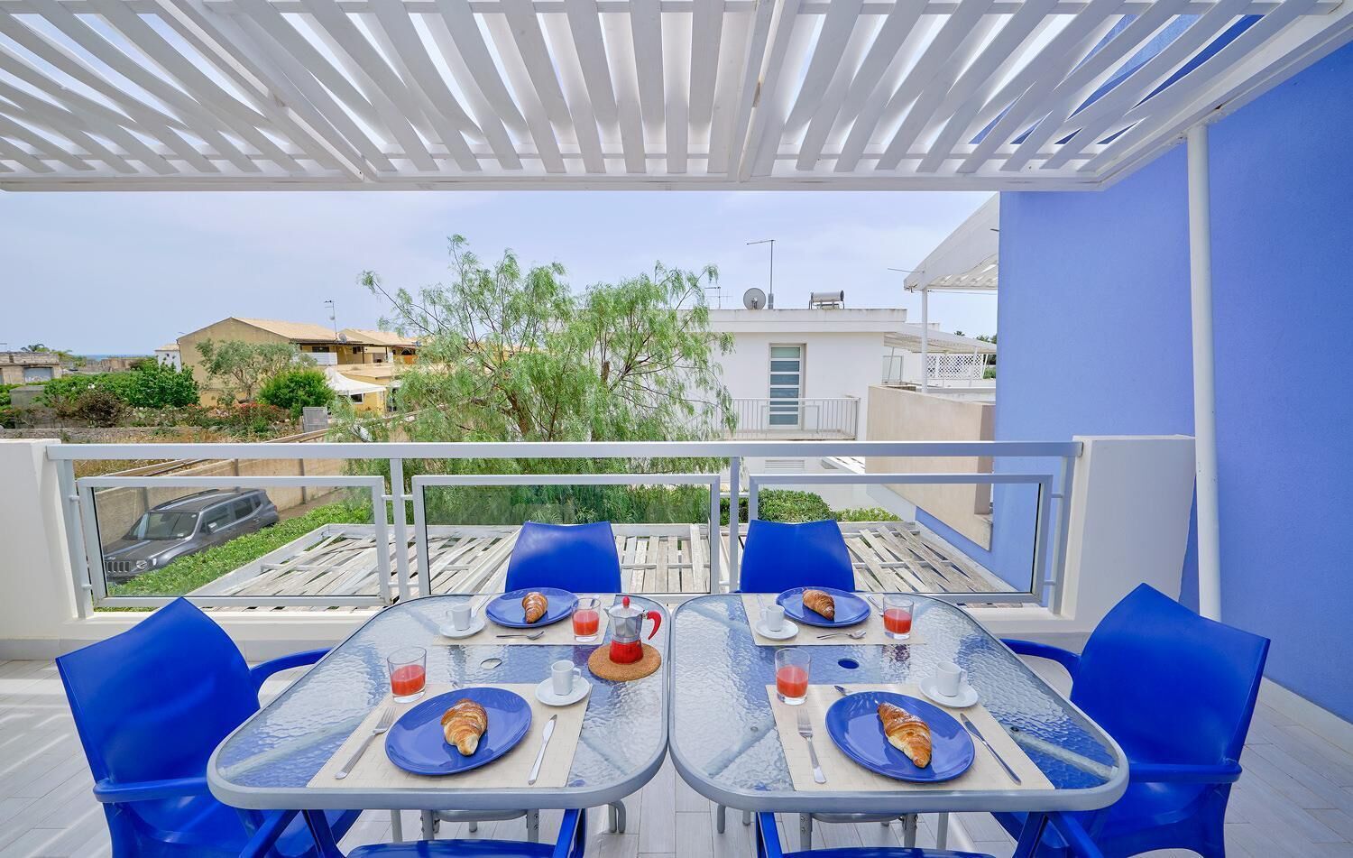 An outdoor dining area with tables and chairs, a view of the surrounding buildings and greenery, and a white pergola overhead.