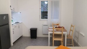 A small, clean room with a kitchenette, a single window, a table with chairs, and a bed with an orange pillow.