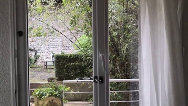 A view through a door with a metal handle, leading to an outdoor area with a stone wall, a bench, and some greenery.