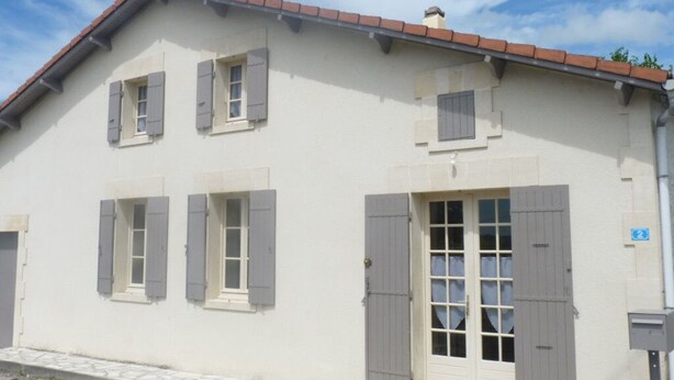 Ein zweistöckiges Gebäude mit heller Fassade, grauen Fensterläden und einem Ziegeldach.
