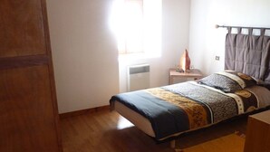 Ein Schlafzimmer mit Holzboden, einem Bett mit gemustertem Überwurf, einem Nachttisch mit Lampe und einem Fenster mit Vorhängen.
