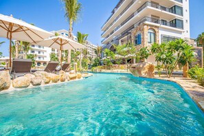 A pool area with lounge chairs and umbrellas, surrounded by palm trees and a multi-story building with balconies.