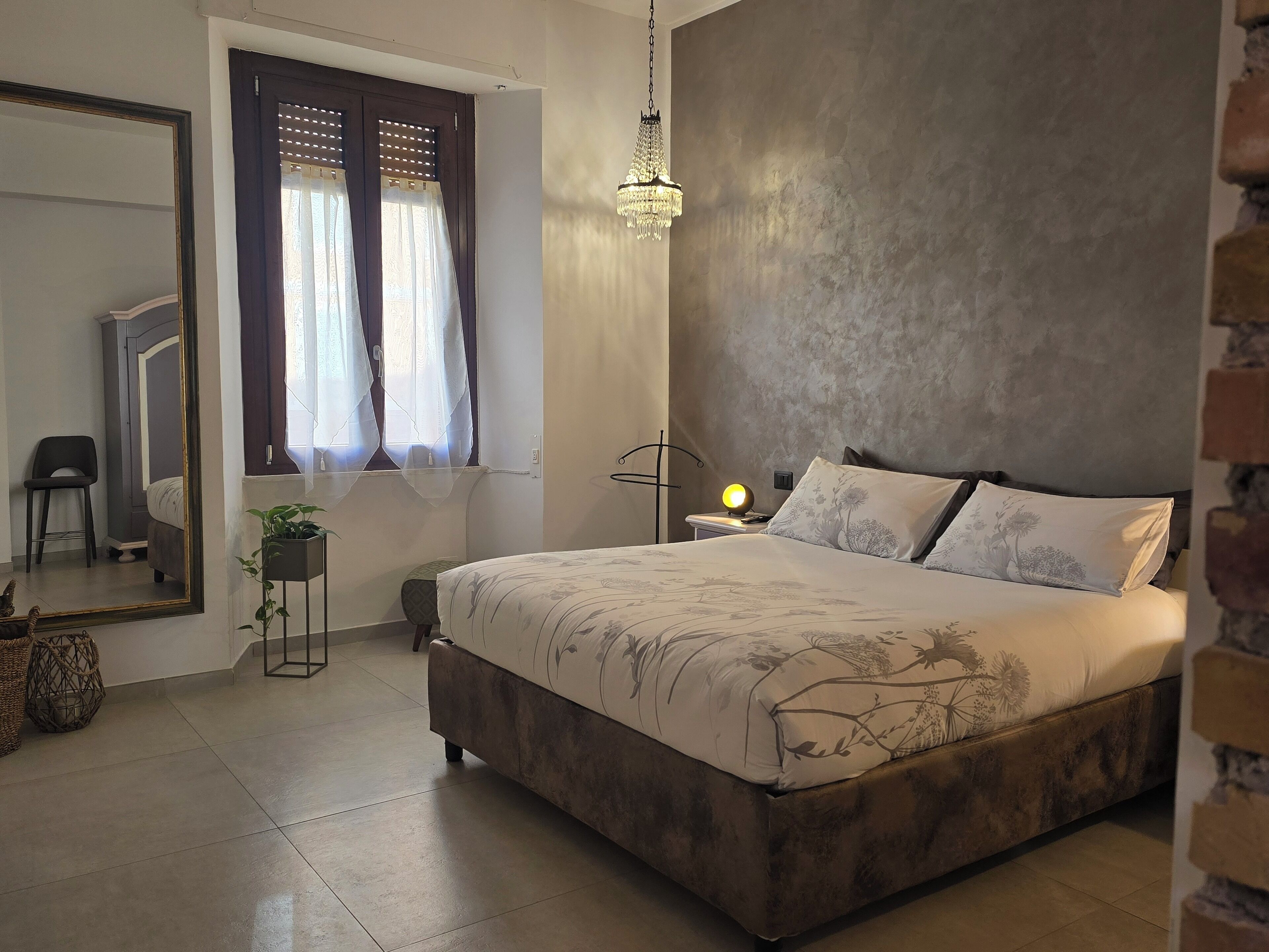 Un dormitorio con cama, mesita de noche con lámpara, espejo, ventana con cortinas y un candelabro colgante.