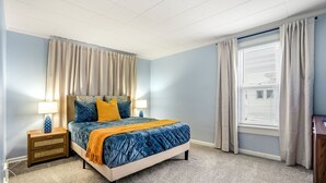 Un dormitorio con cama, mesitas de noche, una ventana con cortinas y suelo alfombrado.