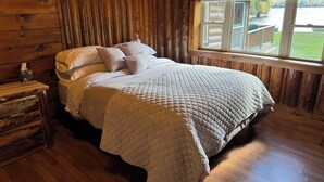 Een houten slaapkamer met een bed, een nachtkastje en een raam met uitzicht op een meer.