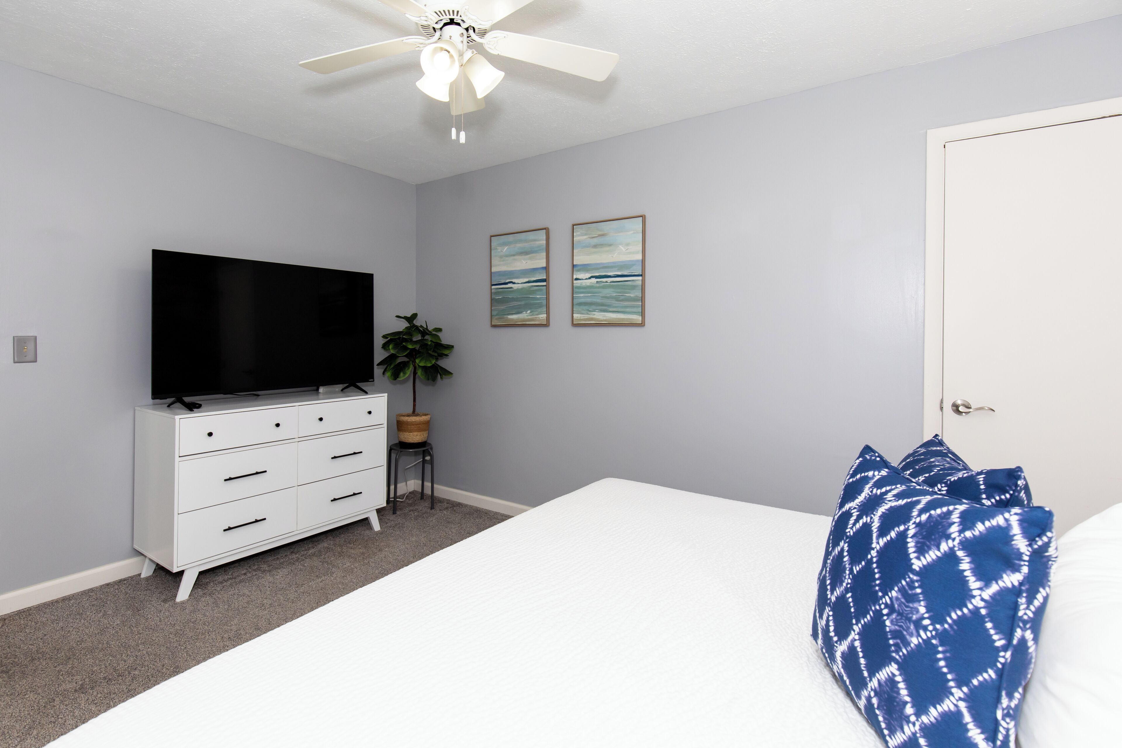 A bedroom with a bed, a TV on a dresser, a ceiling fan, and two framed pictures on the wall.
