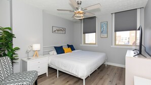 A bedroom with a bed, bedside table, chair, ceiling fan, window with blinds, and a TV.