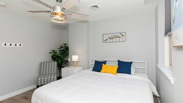 Une chambre à coucher comprenant un lit, un ventilateur de plafond, une table de chevet avec une lampe, une chaise et une étagère fixée au mur.