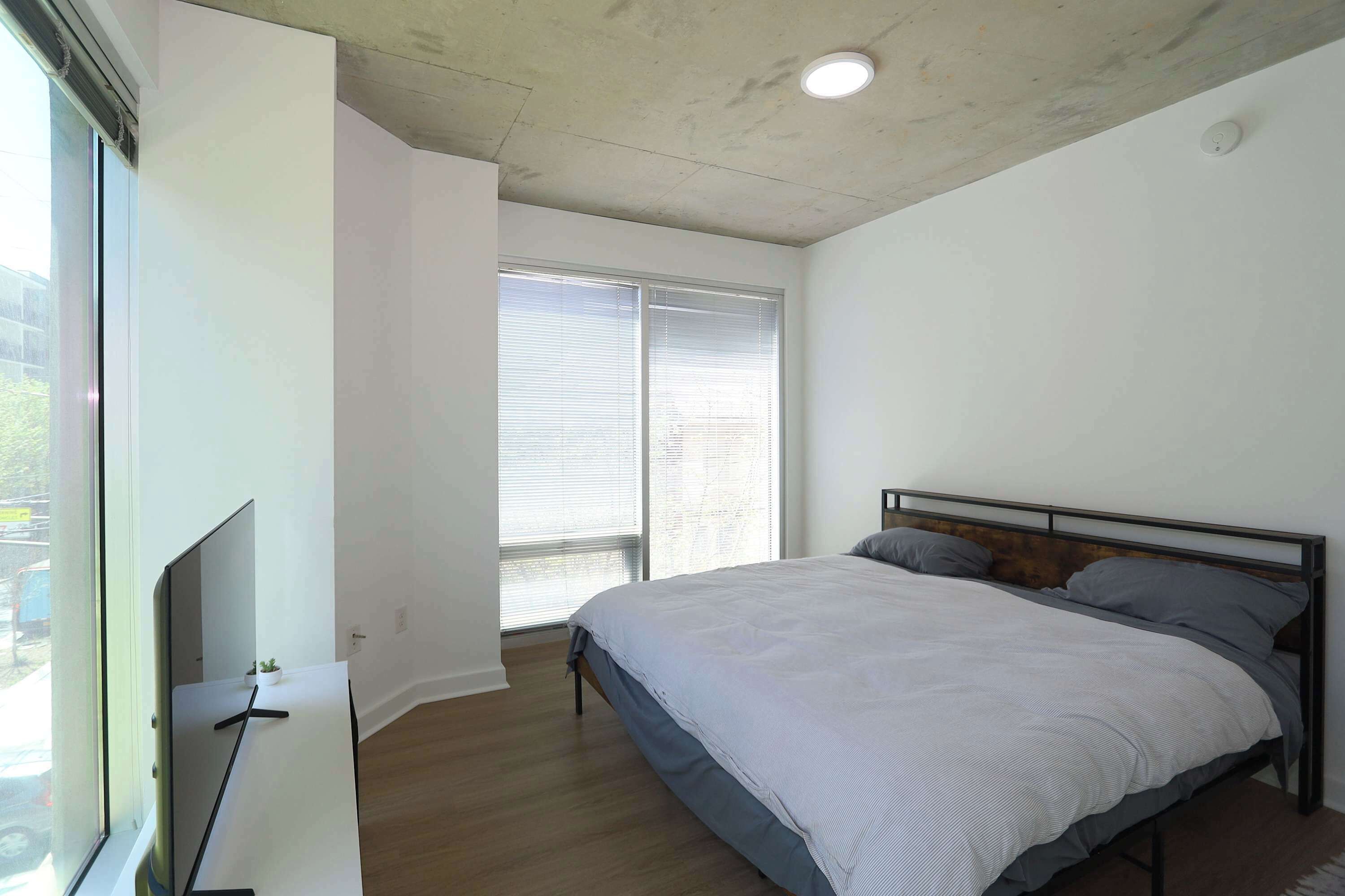 Quarto com cama, televisão, janela com persianas e luminária no teto.