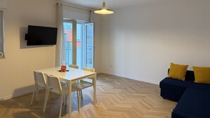 Ein modernes Wohnzimmer mit Essbereich, einem an der Wand befestigten Flachbildfernseher, einer Deckenleuchte und einem Sofa mit gelben Kissen.