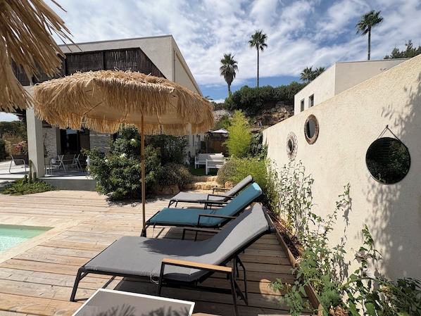 A poolside area with lounge chairs, a thatched umbrella, and a stone wall with circular cutouts.