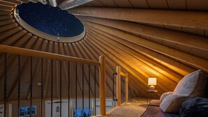 An attic bedroom with a starry view through a circular skylight, a wooden staircase, and a cozy seating area with a lamp. - Stetson + Bronco: 2 Lux Connected Yurts | 5bds 2ba (Canyon Lake)