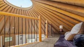 A room with a sloped ceiling, a circular skylight, a wooden railing, a sofa, and a small table with a lamp. - Stetson + Bronco: 2 Lux Connected Yurts | 5bds 2ba (Canyon Lake)