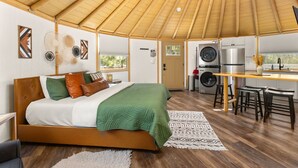 A bedroom with a bed, a wooden ceiling, a kitchen area with a sink, a refrigerator, and a washing machine. - Stetson + Bronco: 2 Lux Connected Yurts | 5bds 2ba (Canyon Lake)